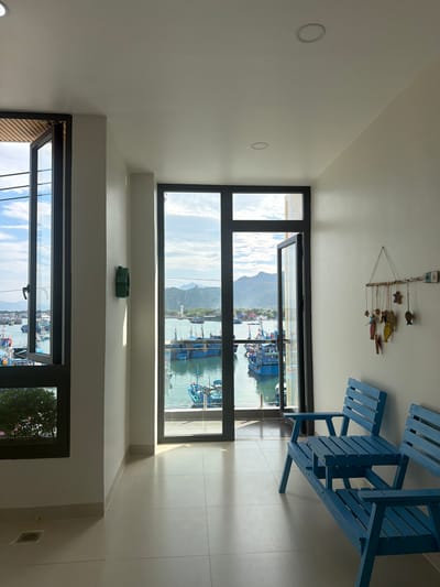 The image displays a brightly lit interior room featuring large, dark-framed windows that offer a clear view of a bustling harbor or bay. The room has light-colored, possibly white or off-white, walls and a light-toned tiled floor.

On the right side of the room, a blue wooden picnic-style table with attached benches is visible, suggesting an indoor or sheltered outdoor dining/seating area. A decorative mobile, crafted from wooden fish and star shapes suspended by strings, hangs from the ceiling. Several recessed circular lights are embedded in the ceiling and upper wall areas. A small, folded green object rests on a white ledge located between sections of the windows.

Beyond the windows, numerous fishing boats of varying sizes and colors are seen in the calm, light blue water of the harbor. Mountains covered in greenery rise in the background under a clear, bright blue sky with scattered white clouds, indicating a sunny day. The scene depicts a coastal environment, aligning with the provided context of Ninh Chu Ward, Vietnam.

No people or direct activities are visible; the room appears to be set up for observation of the outdoor scenery. Near the ceiling where the mobile hangs, a small white rectangular tag is affixed, displaying the text "VEUX 07".