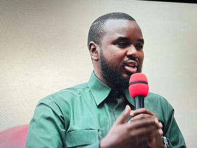 The image shows a Black man with a dark beard speaking into a microphone. He is wearing a green long-sleeved shirt and has a watch on his left wrist. The background appears to be a light-colored wall. The image was taken in Keshero, Democratic Republic of the Congo, at coordinates -1.648 latitude and 29.184 longitude.