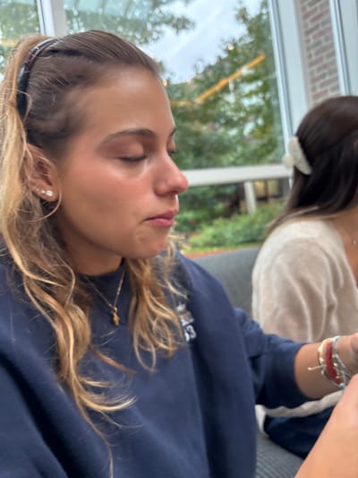The image captures two females seated indoors in Wellesley, United States, next to a large window overlooking an outdoor area. The primary subject, in the foreground on the left, is a young female with light skin, long wavy blonde hair pulled back with a dark hairband. Her eyes are closed, and her mouth is slightly open. She wears multiple stud earrings in her left ear and a gold-colored pendant necklace. Her attire consists of a navy blue crewneck sweatshirt featuring a partially obscured white graphic. Her right arm, visible on the left side of the frame, is adorned with multiple bracelets including red and silver-toned examples.

A second female is partially visible on the right, characterized by dark hair secured with a light-colored claw clip, and wearing a light-colored, possibly beige, long-sleeved top. The immediate indoor environment includes a dark gray upholstered seating surface. The large window reveals an exterior scene with green trees, foliage, and a faintly visible building constructed of light-colored brick or stone. Soft, diffused natural light illuminates the scene. The precise location within Wellesley is not discernable from the visual information present in the image.
