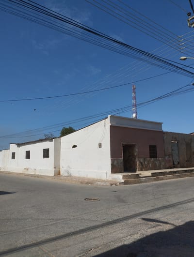The image depicts a street corner in Punta de Piedras, Venezuela. A one-story building occupies the corner, featuring a long white wall with small barred windows and a section with a brown facade, stone accents, and an arched doorway. Adjacent to the brown facade is a decaying structure with an open doorway. The street is paved and shows signs of wear. Overhead, numerous power lines crisscross the sky. In the distance, a telecommunications tower is visible. The sky is clear and blue. There are no people visible in the image.