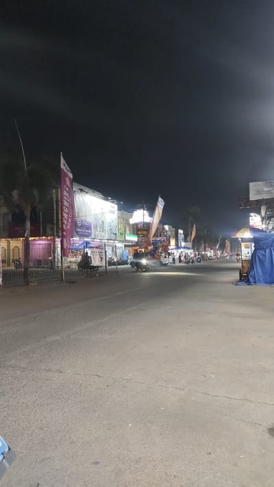 A nighttime street scene in Babelan, Indonesia, shows a road lined with various shops and stalls. The sky is dark, indicating it is nighttime. Several people are visible, some standing and some on motorcycles. The atmosphere appears to be that of a busy commercial area at night, with lights from the establishments illuminating the street.

On the left side of the street, there is a building with a pink facade and windows, possibly a shop or restaurant. In front of it, a metal fence encloses an area. Large vertical banners are displayed, one with the text "LAUNCHING NEW CLUB" and another with the word "Ethica." Further down the street, there are more shops with illuminated signs and displays.

On the right side of the street, there are food stalls or small shops covered with blue tarpaulin. An umbrella is also visible over one of the stalls. Several motorcycles are parked and being ridden on the road, suggesting ongoing activity. A street sign with a yellow arrow is also visible. The road itself is paved and appears to be in decent condition, with some cracks and debris. The lighting creates a vibrant, if somewhat hazy, ambiance typical of a lively night market or commercial street in Southeast Asia.