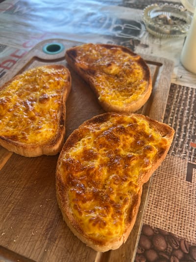 Three slices of toasted bread topped with melted cheese sit on a wooden cutting board. The bread appears golden brown and slightly crispy, with the cheese bubbly and slightly browned in spots. The cutting board has visible scratches and is placed on a table with a patterned surface that resembles burlap and coffee beans. In the background, out of focus, is a glass ashtray with cigarette butts and a glass container filled with a white liquid, possibly milk. The scene is well-lit, with natural light casting subtle shadows. This image was taken in the city of Karabunar, Bulgaria.
