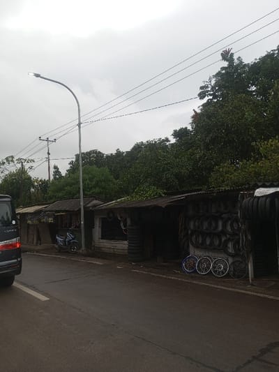 The image depicts a roadside scene in Sukanagalih, Indonesia, under an overcast sky. In the foreground and midground, an asphalt road features a solid white line on its left edge. The rear-left section of a dark-colored minivan is partially visible on the far left. On the right side of the road, a row of single-story commercial structures with corrugated metal roofs extends into the distance. The structure closest to the viewer is a tire shop, displaying numerous black tires stacked and hanging, along with several motorcycle wheel rims, including one blue-colored, leaning against its front facade. Adjacent to it on the left is another shop with visible merchandise inside its open front. A blue scooter is parked facing left on the roadside between these shops and a tall, grey streetlamp with an active LED light, positioned mid-left. Further to the left and rear, a wooden utility pole with multiple cross arms supports overhead power lines that traverse the upper portion of the frame. Dense green trees and foliage form a natural backdrop behind the structures. No individuals are present in the scene.