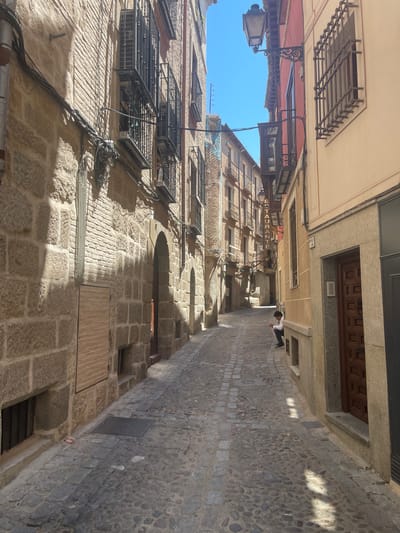 The image depicts a narrow cobblestone street in the historic district of Toledo, Spain. The buildings lining the street are constructed with stone and brick, featuring wrought-iron window grates and balconies. Some of the buildings have a weathered appearance, suggesting their age. The street is illuminated by sunlight, casting distinct shadows on the ground and buildings. A lone individual, a young person wearing a white shirt and dark pants, is seated on a low ledge on the right side of the street, looking down at the ground. The street appears quiet and somewhat deserted. Overhead, a vintage-style street lamp is visible. The architecture suggests a location within a Mediterranean or European city, consistent with Toledo's historical character.