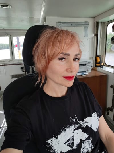 A woman with short, peachy-pink hair and red lipstick is seated in a captain's chair inside what appears to be a boat or ship's cabin. She is wearing a black t-shirt with a white graphic print featuring a hand giving the rock-on sign and a stylized portrait.
The setting includes windows with a view of trees and water, suggesting a location near a river or lake. Inside the cabin, there is a printer on a wooden desk, a life vest, and various equipment mounted on the walls. The lighting suggests it is daytime, and the overall atmosphere is casual. The woman's expression is neutral. There is some visible text on a label on the wall behind her, which reads "VOSS". The location context is Kurdiug, Russia.