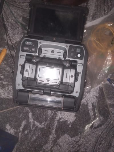 A close-up, high-angle shot shows a fiber optic fusion splicer and its accessories placed on a patterned, dark grey carpet. The fusion splicer is open, revealing its internal components and a small digital display. Control buttons are visible on the top panel of the device, including "SET," "RESET," and "HEAT." A yellow tool, possibly a stripper or cutter, is located to the left of the fusion splicer. To the right of the device, a bundle of yellow fiber optic cables is visible, partially obscured by a clear plastic bag. The overall scene is dimly lit, with some areas of the carpet exhibiting a subtle sheen. The image appears to be taken indoors in Barako, Nigeria. There are no people present in the image.