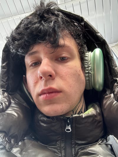 A close-up, eye-level portrait captures a young male subject in Tatishchevo, Russia. The individual, appearing to be in his late teens or early twenties, has dark, curly hair protruding from beneath a dark, glossy puffer jacket hood. His skin is fair, with visible blemishes on the cheeks and forehead. He wears light green over-ear headphones, partially obscured by the hood. Black ink tattoos are visible on the left side of his neck, and a small silver stud earring is present in his left earlobe. The subject maintains a neutral expression, with slightly parted lips, and looks directly at the camera. The background above his head features a light-colored, ridged or slatted structure, indicative of an indoor or covered architectural setting.