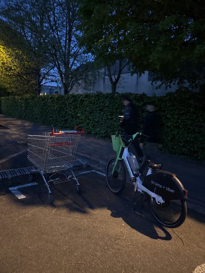 The image, taken at dusk or night in Bures-sur-Yvette, France, depicts a street scene featuring an electric bicycle, a shopping cart, and two individuals.
In the foreground, a metal shopping cart with red handles rests on an asphalt surface. Next to it stands a white and light green electric bicycle, identified by the text "On Lime + Uber" visible on its rear mudguard and "Lime" on its frame. Long shadows from these objects stretch across the pavement, suggesting an artificial light source.
Behind the bicycle and shopping cart, against a backdrop of a wall thickly covered in dark green ivy, two indistinct figures are partially obscured. One figure appears to be holding a small, illuminated item. To the left, a dense canopy of trees with dark foliage is silhouetted against a deep blue, twilight sky. A building with discernible windows is partially visible through the trees and ivy in the background.
The scene is set outdoors on a paved street or path, bordered by lush vegetation. The lighting indicates either early evening or night, with some artificial illumination highlighting the foreground objects and ground, creating strong shadows and a generally dim atmosphere. There is no clear dynamic activity taking place, rather a static arrangement of objects and figures in a quiet, nocturnal setting.
