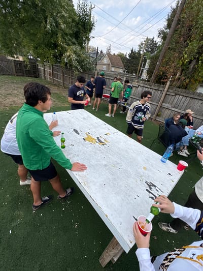 The image depicts an outdoor social gathering in the backyard of a residential property in South Bend, Indiana, United States. The ground is covered with artificial green turf. A large, weathered white rectangular table, exhibiting significant paint peeling and exposed yellow and dark areas, is centrally positioned.
Approximately 10-12 individuals, predominantly young adults, are present. In the foreground, a male wearing a green long-sleeved shirt and black shorts stands next to the table, holding a clear plastic water bottle and a green glass bottle. To the right, another male, wearing a white long-sleeved shirt and a navy and beige Notre Dame hockey jersey, is actively pouring liquid from a green glass bottle into a red plastic cup held in his left hand. Additional red plastic cups and a green glass bottle are on the table.
In the mid-ground, a male wearing a navy "Notre Dame" t-shirt is opening a red beverage can near a red cooler on the ground. A group of other males, some wearing green attire, are standing around the cooler and a small black-and-silver striped object. In the right background, two females are seated on black metal outdoor chairs; one wears a straw hat.
The background features a wooden fence, mature trees with sparse foliage, and residential houses under an overcast sky. Overhead utility lines and wooden power poles are also visible. The scene suggests a casual party or tailgate event, with several participants wearing apparel associated with the University of Notre Dame.