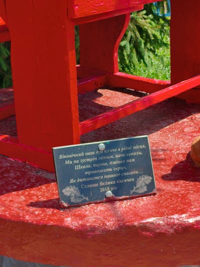 A dark blue plaque with white text is affixed to a bright red, possibly painted, wooden structure. The text on the plaque appears to be in Ukrainian and reads: "Віковічний наш дуб кличе в рідні місця, Ми на зустріч летим, наче соколи, Школа, парта, дзвінок нам тривожать серця, Як дитинства нашого спогади... Селище Велика Багачка 2018 рік". This translates to: "Our eternal oak calls to native places, We fly to meet, like falcons, School, desk, bell disturb our hearts, Like our childhood memories... Village of Velyka Bahachka, 2018." The structure itself is painted a vivid red and is situated outdoors, with blurred green foliage visible in the background, suggesting a natural or park-like setting. The plaque has a metallic screw or fastener visible on its left side.