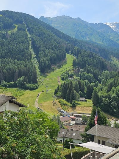 This is an eye-level, wide-angle, full shot of a mountainous landscape in Pontagna, Italy, on a clear, sunny day. In the foreground, a white canopy-covered outdoor dining area is visible with a table and chairs. To the right of the canopy, a residential building with a grey tiled roof and an American flag flying from a flagpole are present. In the midground, more residential buildings with grey tiled roofs are scattered among lush green trees. A ski lift with numerous chairs ascends the grassy mountainside, surrounded by dense evergreen trees. Further up the mountain, a cleared ski slope is visible. In the background, majestic, rugged mountains with snow-capped peaks dominate the skyline.
