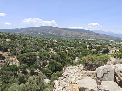 This is a wide-angle, eye-level shot of a hilly, arid landscape under a bright blue sky with scattered white clouds. In the foreground, large, light-colored rocks are prominent. The middle ground is densely populated with olive trees and other green vegetation, interspersed with dry, rocky terrain. In the mid-distance, a partially ruined stone structure, possibly an old tower or chimney, is visible amidst the trees. The background features a large, undulating mountain range covered in sparse vegetation and some scattered buildings. In the far distance, lighter blue mountains are visible. Wind turbines are present on the distant mountain crests. The scene appears to be located in a rural area, possibly on the outskirts of Ierapetra, Greece, known for its olive groves and mountainous terrain. There are no people or animals visible in the image.