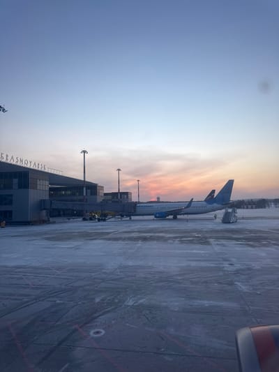 An outdoor wide-angle photograph taken at Емельяново (аэропорт) in Krasnoyarsk, Russia, depicts an airport apron scene during sunrise or sunset, characterized by a blue, pink, and orange sky. The foreground, from the perspective of an aircraft window, reveals a portion of a red and blue engine nacelle. The mid-ground features a snow-dusted concrete apron with tire tracks and red demarcation lines. A white and light blue Pobeda Airlines aircraft, bearing the word "ПОБЕДА" on its fuselage, is parked at a gate, connected to a jet bridge extending from the terminal building. A mobile boarding stair is positioned near the aircraft's rear passenger door, and one individual in a high-visibility vest is visible on the apron near the aircraft. To the left, the modern, multi-story airport terminal building is visible, with the word "KRASNOYARSK" displayed prominently on its roofline. Several tall airport light poles punctuate the scene. In the background, an air traffic control tower stands against the colorful sky, with a distant treeline and snow-covered terrain completing the horizon.