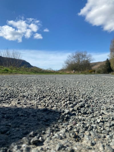 The image depicts a low-angle, perspective view of an asphalt road or pavement surface, textured with small aggregate stones, in Llangynog, United Kingdom. The road occupies the left half of the frame. The right half shows a landscape comprising grassy areas, shrubs, trees, and gently sloping hills. A blue sky with scattered white cumulus clouds fills the upper portion of the scene.