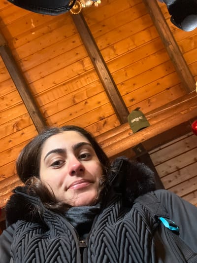 A young adult female is depicted in a low-angle selfie, centered in the lower half of the frame, looking directly at the camera with a slight smile. She has dark hair and fair skin. She is wearing a black puffer jacket with a textured, chevron-patterned front panel, a dark fur-lined hood, and a dark turtleneck garment underneath. A black backpack strap with a light blue buckle is visible on her right shoulder.
The background consists of a warm-toned wooden ceiling, composed of horizontal planks and two diagonal dark wooden beams. An olive green baseball cap with the white text "APRÈS SKI" is hanging on the rightmost beam. In the upper left corner, a partial view of a golden, ornate fixture is visible. A red spherical ornament is partially visible in the upper right corner.
The scene takes place indoors, likely within a rustic-style building in the city of Valtournenche, Italy.