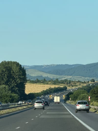 The image depicts a highway scene in Dolni Bogrov, Bulgaria, on a clear, sunny day. The highway has multiple lanes and is populated with various vehicles, including cars and trucks. In the foreground, a silver car is driving on the right lane. A large green tree is visible on the left side of the image, with a field of dry grass behind it. In the background, rolling hills and mountains are visible under a clear blue sky. The overall impression is a busy highway cutting through a rural landscape.