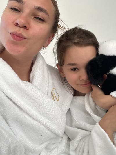 Two individuals, likely an adult and a child, are pictured in a close-up shot, both wearing white terry cloth robes. The adult, on the left, is looking towards the camera with their lips pursed. The child, on the right, is looking slightly away from the camera and holding a black and white plush toy. The robes appear to be soft and absorbent, suitable for spa or bath use. The background is a plain white wall, suggesting an indoor setting. The image quality is high, with clear focus and good lighting. The scene does not provide any specific clues about the location in Terskol, Russia.