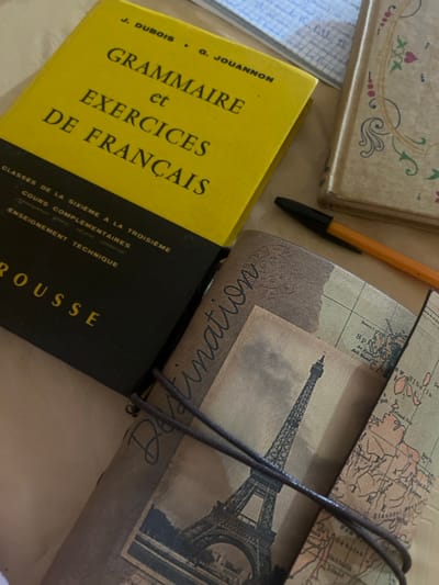 In Ksar-el-Kebir, Morocco, a collection of academic and personal items is arranged on a light-colored, possibly beige, surface, viewed from an overhead perspective. Dominating the upper-left quadrant is a used, yellow and black hardback textbook titled "GRAMMAIRE et EXERCICES DE FRANÇAIS" (Grammar and French Exercises) by J. DUBOIS and G. JOUANNON, published by LAROUSSE, indicated for "CLASSES DE LA SIXIÈME A LA TROISIÈME" and "COURS COMPLÉMENTAIRES" including "ENSEIGNEMENT TECHNIQUE". Adjacent to its right, partially visible, is a decorative beige or light-brown journal with a spine featuring intricate red, green, and black floral or vine patterns.

Below these, slightly off-center, lies a brown traveler's notebook or journal with a textured cover. The cover displays the word "Destination" in cursive script, an aged sepia-toned image of the Eiffel Tower, and a vintage-style world map. A dark brown elastic cord secures the notebook. A black and orange ballpoint pen rests diagonally between the decorative journal and the traveler's notebook, its tip pointing towards the Eiffel Tower image. In the upper-right background, a sheet of light blue lined paper from a notebook is visible, displaying faint handwritten text. No individuals are present in the scene. The items suggest an environment related to study, travel planning, or personal documentation.