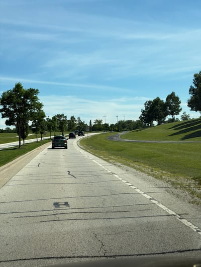 The image captures a view of a curved road in Vernon Hills, United States of America, on a bright, sunny day with a blue sky and wispy clouds. The road is paved and has white lane markings. Several cars are visible on the road, driving in the same direction. On the left side of the road, there are mowed grass areas and several trees with green foliage. On the right side of the road, a grassy hill slopes upwards, dotted with trees that cast shadows on the grass. In the distance, a few more trees and some structures, possibly lights for a sports field, can be seen. The overall scene depicts a suburban or park-like environment with a roadway winding through a green landscape.