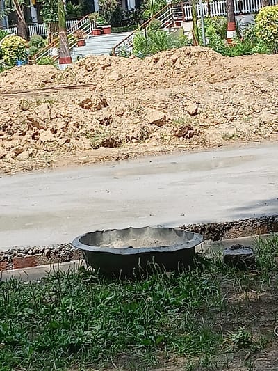 The image shows a large, round, black planter filled with sand and sitting on a paved area in front of a large pile of dirt. The planter is partially obscured by tall green grass. The paved area is bordered by a small, dirt berm and there is a brick visible in the dirt. In the distance, a set of stairs leads up to a white fence. The image appears to have been taken in Dehradun, India, at the coordinates (30.304, 78.017).