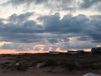 The image shows a sunset scene in Blagoveshchenskaya, Russia. The sky is filled with dramatic, dark clouds with streaks of orange and pink peeking through from the setting sun. In the foreground, there is a sandy, desolate landscape with scrubby vegetation and some scattered debris, possibly driftwood. In the middle ground, there are several buildings, some appearing to be modern residential or commercial structures, with flat roofs. The overall atmosphere is moody and serene. There are no people visible in the image.