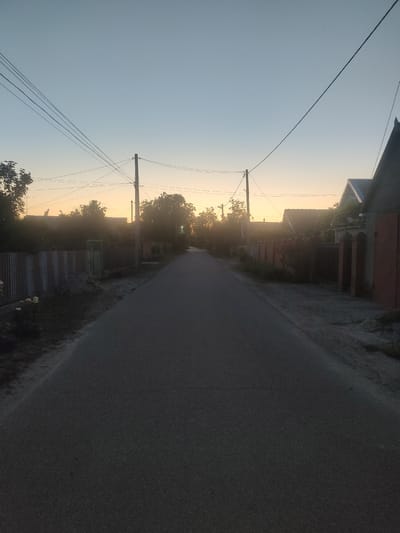 The image depicts a rural street scene at dusk, likely in Ternovka, Moldova. A paved road runs through the center of the frame, disappearing into the distance. On either side of the road are residential properties with fences and houses. Power lines are visible crisscrossing the sky, supported by wooden poles. The sky is a gradient of pale blue and orange, indicating the setting sun. There are trees and some vegetation along the roadside. The overall impression is one of quiet evening in a village setting.