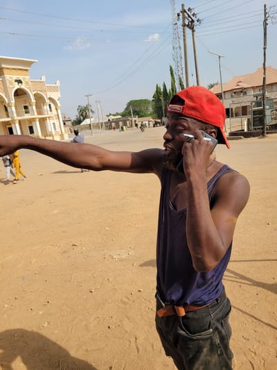 A dark-skinned male is centered in the image, wearing a red baseball cap with "49ERS" visible on the back, a dark blue sleeveless tank top, and dark trousers with a brown belt. He holds a dark-colored smartphone to his right ear, appearing to speak, with a small light-colored cylindrical object lodged between the phone and his ear. His left arm is extended straight out to the left, with the hand seemingly pointing. He stands on a light brown, sandy or dusty ground with scattered small rocks and debris. His shadow extends from the lower-left.

In the background to the left, a prominent two-story building with beige walls, ornate golden trim, arched windows, and white columns is visible. An emblem, possibly an elephant head, is present above an arch on the upper floor. Further left, two individuals walk on the dirt ground, one dressed in a yellow garment.

To the right, two utility poles with numerous overhead wires stand against a clear blue sky with minimal cloud cover. A metal lattice communication tower is visible in the far distance. Further right, a building with a reddish-brown tiled roof and light-colored walls is partially obscured by fencing topped with barbed wire. The general environment is an outdoor street or open area in Nassarawa Egon, Nigeria, characterized by dry, sandy earth, diverse architecture ranging from ornate to simpler structures, and visible utility infrastructure including power lines and a cell tower. Distant figures and what appears to be a motorcycle are visible further down the road.