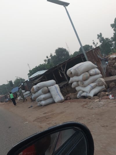 Sacks line a roadside alongside a makeshift stall. The ground is rough and appears unpaved. A utility pole with a solar panel stands nearby. A car's side mirror is visible in the foreground, suggesting the photo was taken from inside a vehicle. In the distance, trees and a tower-like structure can be seen through the hazy atmosphere.