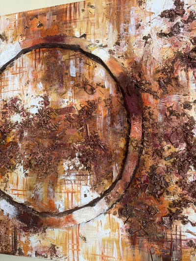 This is a close-up, high-angle, eye-level shot of an abstract painting with a textured surface. The painting features a prominent circular form outlined in dark brown with a white textured band on the inner circumference. The background is a chaotic blend of orange, white, and brown brushstrokes, with visible texture from what appears to be wood shavings or dried organic material applied to the canvas. There are no people, animals, or recognizable landmarks present in the image. The image does not depict any location in the city of Dodoma, Tanzania.