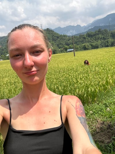 A selfie, taken in portrait orientation but displayed rotated, captures a young adult female in a vast natural setting under a partly cloudy sky. The main subject, positioned in the foreground, looks directly at the camera with a faint smile. Her fair skin appears slightly reddened from sun exposure, and her light hair is pulled back. She wears a black strap top, revealing her left shoulder and arm, which bears a vibrant, abstract tattoo in shades of orange, blue, and black. Several moles are visible on her chest and shoulder.
Behind her, an expansive field of tall, vibrant green crops, likely rice, stretches into the middle ground. Partially obscured by the crops, the head and shoulders of a second person are visible in the middle distance. A wooden pole with a small, unreadable blue sign is also present in the field.
The background features multiple layers of majestic mountains and hills, densely covered in green vegetation. The most distant mountains appear hazy and blue against a sky with scattered white and gray clouds. The bright sunlight suggests it is late morning or afternoon. The scene conveys a peaceful, rural atmosphere, consistent with locations like Ngọc Đường Commune, Vietnam. No readable text is visible on the small blue sign.