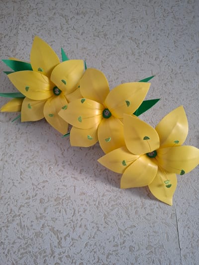 Three artificial yellow lilies with green leaves and stamen with glitter accents are arranged diagonally on a textured, light-colored surface. The lilies are constructed from a foam-like material. The background surface is off-white and has a raised, patterned texture with some shiny flecks. The image is taken from a slightly elevated angle, looking down at the lilies. There are no people visible in the image. The setting does not provide any information to determine a specific location within Jitiqara, Kazakhstan.