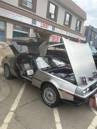 A silver DeLorean DMC-12 sports car with its gull-wing doors and hood open is parked on an asphalt street in front of a two-story building with a brick lower facade and wood-paneled upper facade. The building has signs indicating "Housekeeping Award," "WALDORF HOTEL," "LOBBY," and "Chinese Western Cuisine Waldorf Restaurant." The DeLorean's interior is visible, showing gray leather seats and a "Keep Your DeLorean Stainless Steel CAR CARE KIT" box. The engine compartment is open, revealing various items and products. Several Funko Pop figures and other merchandise are displayed on the edge of the opened hood. A Canadian flag is attached to the building's exterior above the restaurant sign. The street is marked with white lines, and there are some cracks and stains on the asphalt. Other vehicles and a lamppost are visible in the background. The scene appears to be an outdoor event or car show in the City of Leduc, Alberta, Canada, based on the context of the "WALDORF HOTEL" and nearby businesses.