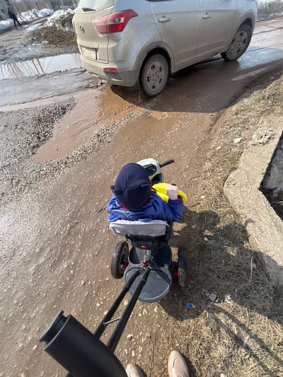 The image depicts a rural or semi-urban outdoor scene in Gamovo, Russia, under bright daylight conditions, likely during a spring thaw. An adult, whose light-colored shoes are visible in the foreground, is pushing a black and grey child's tricycle with a black push handle. Seated in the tricycle is a small child, viewed from above and behind, wearing a dark blue hat with ear-like features and a blue jacket, holding yellow handlebars.
The ground consists of a heavily degraded unpaved road or path, covered extensively with wet, dark brown mud and gravel. Numerous large puddles of muddy water reflect the sunlight. A silver Hyundai Creta SUV, noticeably dirty, is parked or slowly moving in the midground, partially submerged in a large puddle. The vehicle's rear license plate is visible, displaying "M157BE 92". To the right, a concrete curb separates the muddy path from an area of dry, light brown grass. In the far background, beyond the vehicle and more mud puddles, patches of dirty snow or ice are visible, along with bare trees and a distant, blurred human figure.