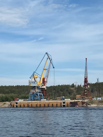 The image depicts a waterfront scene in Nizhny Bestyakh, Russia, featuring industrial equipment along the shore of a wide river. In the foreground, dark blue water with visible ripples occupies the lower portion of the frame. Dominating the mid-ground is a large, tan-colored concrete structure, likely a dock or pier, with multiple cylindrical fenders attached along its edge. Several shipping containers and other industrial materials are visible on top of this structure.
Two prominent cranes stand on the dock. The larger crane on the left is a multi-colored gantry crane with a blue base, a yellow boom, and a red counterweight section. Its long boom extends upwards and to the right. To the right of this crane is a taller, slender red lattice boom crane, positioned further back on the shore. Its boom is oriented vertically.
Behind the dock and cranes, a dense line of dark green trees forms a backdrop, suggesting a forested area. The sky above is a bright blue with scattered white clouds. In the distance, on the right side of the image, a small structure with a tall pole or antenna is visible. The overall impression is of an active industrial port or shipyard. No people are discernible in the image.