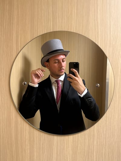 A male individual is depicted in a mirror selfie, standing in an indoor setting with light-colored, vertically-grained wooden wall paneling serving as the primary background. He is formally dressed in a dark suit jacket worn over a white dress shirt. A reddish-pink tie, featuring small, repeating animal motifs that appear to be horses, is knotted around his neck. On his head, he wears a grey top hat with a distinct dark band, which he is subtly adjusting with his left hand by touching the brim. His right hand holds a black smartphone, equipped with multiple rear cameras, positioned to capture his reflection. The man has short dark hair visible beneath the hat and a gold hoop earring in his left earlobe (as seen in the mirror). His expression is focused, looking downwards. A small, circular, metallic fixture is visible on the wooden wall, reflected in the mirror to the left of the man. The formal attire, specifically the top hat and the tie's pattern, strongly suggests preparation for a formal event such as the Epsom Derby, consistent with the specified location of Epsom and Ewell, United Kingdom.