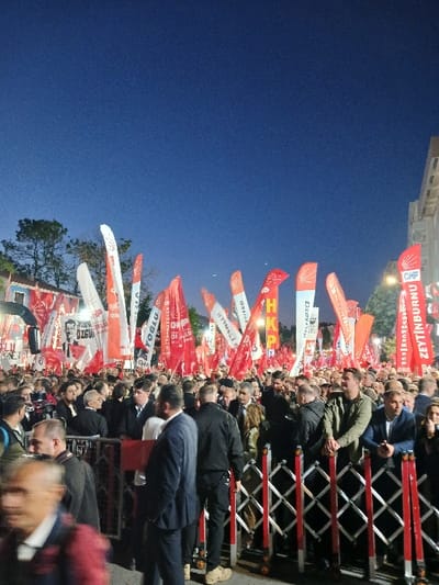 A large crowd of people, mostly men, are gathered in front of a metal fence barrier, facing red and white flags. The flags are predominantly red, with some white ones interspersed. The crowd is densely packed, and the individuals are mostly obscured by the flags and the sheer number of people. The scene is lit by the light of street lamps and ambient city lights. The sky is a dark blue, with a few visible stars. The event is taking place in Silivri, Türkiye, at the location indicated by coordinates 41.071, 28.244. The crowd appears to be attending a political rally or a public event. The people are generally dressed casually, and there are no visible signs of conflict or tension. The atmosphere seems to be celebratory, with flags and banners, suggesting a gathering of supporters for a cause.