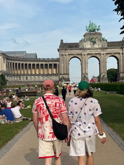 The image shows two individuals walking on a paved path in a park in Brussels, Belgium, near the Cinquantenaire Arch (latitude: 50.841, longitude: 4.389). The person on the left is wearing a red and white patterned shirt with a black crossbody bag, beige shorts, and a red baseball cap. The person on the right is wearing a white shirt with blue floral patterns, white shorts, and a green baseball cap. They are walking toward the arch, which is a large, ornate structure with three arches. The arch is surrounded by trees and greenery, and there are people sitting on the grass and benches in the background. The sky is blue with a few white clouds. The scene is peaceful and sunny.