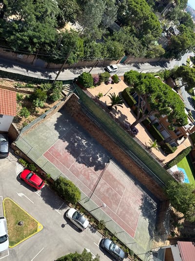 This aerial shot captures a sunny day in Sant Feliu De Guíxols, Spain, showcasing a residential area with a tennis court and a road. The scene is dominated by lush green trees and vegetation, suggesting a warm, pleasant climate.
The focal point of the image is a tennis court with a red and green surface, surrounded by a fence. Shadows from trees are cast across the court, indicating it is likely daytime, possibly mid-morning or afternoon. Adjacent to the tennis court is a swimming pool with a blue cover, hinting at recreational activities.
A winding road passes through the scene, with several cars parked along its edge and in a small parking area. These include a white hatchback, a red hatchback, and a dark-colored sedan. The buildings visible are residential, with terracotta roof tiles and beige facades.
The overall impression is one of a peaceful, well-maintained residential area in a sunny Mediterranean location. There is no visible text in the image.