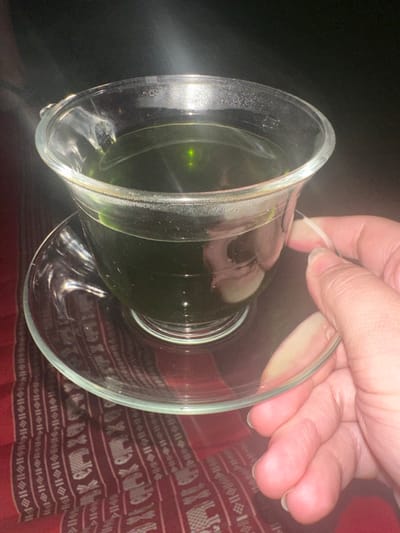 The image depicts a close-up, elevated perspective of a clear glass teacup and saucer, held by a human hand, in the city of Ban Paem Bok, Thailand. The teacup contains a dark green liquid, filling approximately two-thirds of its volume, with a distinct bright green light reflection visible on the liquid's surface. A light-skinned human hand, positioned on the right side of the frame, uses its thumb and index finger to grip the rim of the glass saucer. The glass objects are situated above a dark red textile surface that features repetitive patterned designs, including motifs resembling elephants and linear stripe arrangements in a lighter color. The background is dark and indistinct.