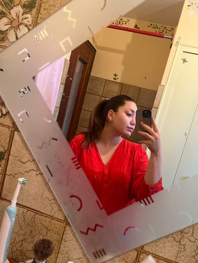 The image displays a female individual taking a mirror selfie in a bathroom, identified as being in Ancenis, France. The subject is reflected from the chest up, holding a dark-colored smartphone in her left hand (appearing as her right in the reflection), positioned to capture the image. She has light brown hair styled in a ponytail and is wearing a red, long-sleeved, V-neck top with front buttons and eyelet embroidery.
The mirror features a frosted frame with geometric cutouts including zigzags, straight lines, rectangles, and semicircles; small water droplets are present on the mirror's surface. The reflected background reveals a bathroom interior: the lower wall section is covered with brown, textured square tiles, while the upper wall is painted a light cream color. Decorative elements on the upper wall include a red border strip with a leaf pattern and several small green frog decals.
On a surface to the left of the subject's reflection, a light blue and white electric toothbrush and a shaving brush with a dark handle are visible. Further within the reflection, a dark wooden door with a vertical glass panel and a light pink towel are discernible. A light-colored cabinet or door is visible to the right of the subject's reflection.