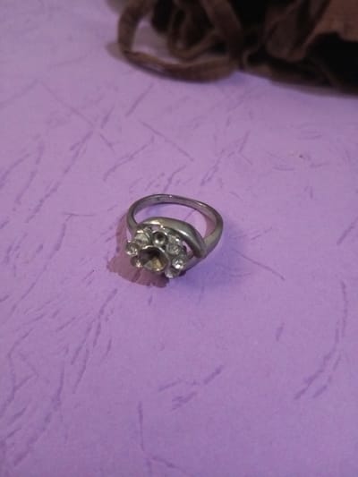 A silver-colored ring is positioned slightly off-center on a textured, light purple surface. The ring features a top cluster design: a central circular setting, which appears dark or empty, surrounded by seven smaller, clear, faceted stones arranged in a floral or circular pattern. The metal band exhibits a slightly dull, possibly worn, finish. A blurred, dark brown, amorphous object is visible in the upper background. No individuals, actions, or interactions are present. The scene is located in the city of Chandragiri, Nepal.