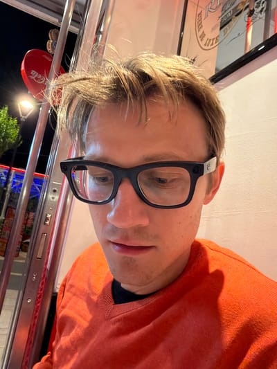 A selfie, rotated, shows a man with blond hair, wearing black-rimmed glasses and an orange sweater. He is seated at a table with a silver metal frame, possibly in a diner or restaurant. The table is partially visible, with a red Coca-Cola branded object hanging nearby. Through a window, there's a partial view of a tree and night sky, with lights suggesting an urban environment. Based on contextual clues, the location is Ragusa, Italy. A framed image on the wall is partially visible.