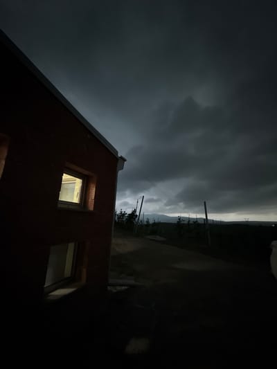 A nighttime exterior scene in Sisian, Armenia, features a red brick building on the left with two illuminated windows. A dirt road leads away from the building towards a dark, hilly landscape under a heavily clouded, stormy sky. Several wooden poles and some sparse vegetation are visible along the road. A power line stretches across the sky. The overall mood is dark and somber, suggesting approaching bad weather.