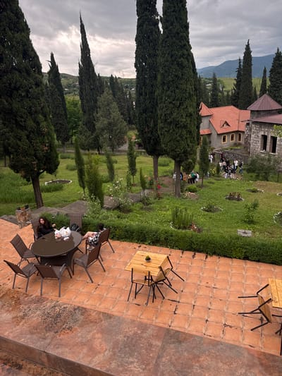 In Akhtala, Armenia, a scenic overview captures a multi-story stone building with a red-tiled roof, surrounded by lush greenery and tall cypress trees under a cloudy sky. In the foreground, an outdoor patio area features several tables and chairs. Two individuals are seated at a round table, engaged in conversation. Further into the garden, a group of people are observed walking together. The environment suggests a tranquil, possibly historical or resort-like setting.