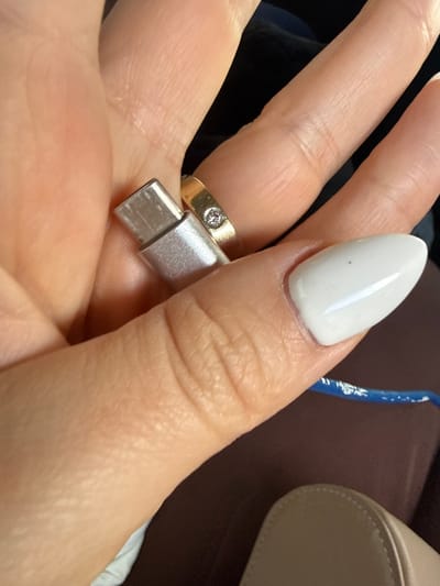 An extreme close-up shot focuses on a person's left hand holding a silver-colored USB-C connector. The hand exhibits light-toned skin with visible wrinkles and knuckle lines. A gold-colored band ring, set with a single round clear stone, is visible on the ring finger. The thumb, positioned to the right, has an elongated, oval-shaped fingernail coated in opaque white polish, featuring a small dark speck on its surface. The USB-C connector is oriented vertically, held between the thumb and the base of the index or middle finger. In the lower right background, a blurred segment of a blue cable with white markings is visible, alongside a curved edge of a light-colored, possibly beige or cream-colored, object, which appears to be part of a shoe or bag. The immediate environment is dimly lit and out of focus, suggesting an indoor or vehicle setting. The specific location in Apnagyugh, Armenia, cannot be determined from the image.