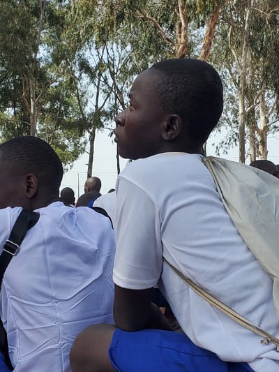 The image shows a medium shot of a group of young people, likely students, from the chest up, seated outdoors. The main subject, a young person with dark skin and short hair, is positioned in the foreground, facing to the right. They are wearing a white t-shirt with blue shorts. A light-colored cloth is draped over their left shoulder, fastened with a zipper-like detail. Behind them, other individuals are also visible, mostly wearing white tops. The background consists of tall trees with sparse foliage, suggesting a rural or semi-rural environment, possibly within Barakin Rafin Gora, Nigeria. The lighting indicates it is daytime, with the sun casting soft shadows. The overall atmosphere appears calm and focused, as if the subjects are observing something or waiting for an event.
