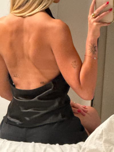 An indoor scene in Alfei Menashe, Palestinian Territory, depicts a female subject from the back and right side, seated. The subject has blonde hair, partially visible over the right shoulder, and tan skin. She is wearing a black, backless garment, possibly made of satin, which exposes her upper back. Several tattoos are visible: faint script on the left lower back, three small vertical lines on the right lower back, a circular symbol on the right upper arm, and a distinct sun and crescent moon design on the right forearm. The subject's right hand, adorned with red nail polish and a gold-toned ring on the ring finger, is holding a light-colored object, likely a smartphone or reflecting surface, suggesting a self-portrait. A thin bracelet with a small, light-colored bead is visible on her right wrist. Her left hand, also with red nail polish, rests on her right thigh. The immediate foreground shows a white, textured surface, possibly a bed or cushion, indicating the subject is seated. The background is an indistinct, light-colored wall or interior surface.