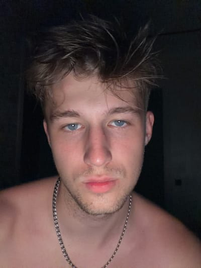 A young adult male with light brown hair and blue eyes is looking directly at the camera. He has a silver chain around his neck. The background is dark and indistinct, suggesting an indoor environment. There are no other people or objects clearly visible in the frame. The image is a close-up portrait, focusing on the man's face and upper chest.