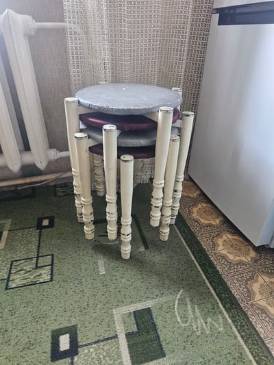 A stack of three small stools with turned wooden legs painted white with distressed details sits on a green carpet with a geometric pattern. The stools have round, padded seats covered in gray and dark purple fabric. To the left, a white radiator is attached to a beige, textured wall. Behind the stools, a white, net-like curtain hangs. To the right of the stools, a portion of a white refrigerator is visible. The floor in front of the refrigerator is tiled with an ornate beige and brown pattern. The scene is indoors, likely in a residential setting.