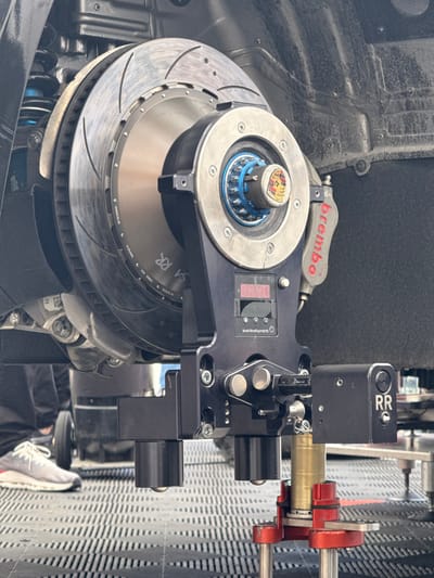 The image shows a close-up of a black car's rear right wheel, which is being inspected with a black device attached to the caliper. The device is labeled "Invel Developments" and has two cylindrical arms, one labeled "RR". The car is on a black platform with a grid pattern. The brake caliper is labeled "Brembo" and has a red, white, and blue sticker with the Porsche logo. The image is taken in Stavelot, Belgium, at coordinates 50.441 latitude and 5.969 longitude. A pair of sneakers, partially visible, are in the foreground. The photo was taken at a car racing event, likely at the Spa-Francorchamps racetrack, near the town of Stavelot.